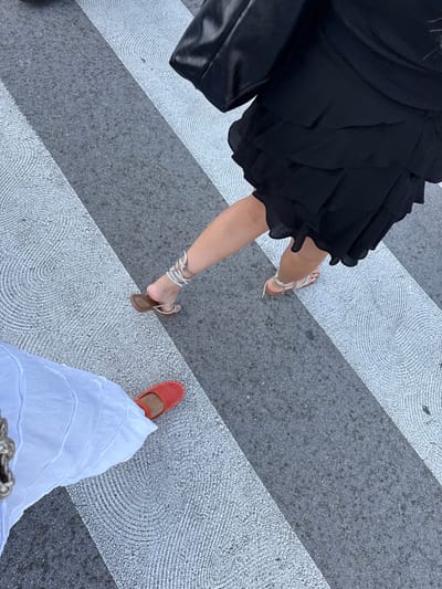 The image presents an overhead, slightly angled view of two individuals crossing a street in Santander, Spain. The ground is a pedestrian crossing composed of alternating dark grey asphalt and light grey painted stripes, with the lighter stripes featuring a debossed or textured pattern of concentric circles.
One individual, positioned on the right and extending from the upper-middle to the lower-right, is wearing a black, layered, ruffled skirt or dress. This person wears light-colored, multi-strapped sandals, with straps wrapping around the lower leg/ankle. A portion of a black handbag is visible in the upper-right corner.
The second individual, visible in the lower-left, is wearing white, textured, wide-leg trousers or a long skirt. This person wears bright orange, espadrille-style flat shoes. A braided, metallic-toned strap, possibly from a bag, is partially visible in the far upper-left corner, near this person's leg.
Both individuals are in motion, positioned on the pedestrian crossing, suggesting they are either walking concurrently or in close proximity across the street.