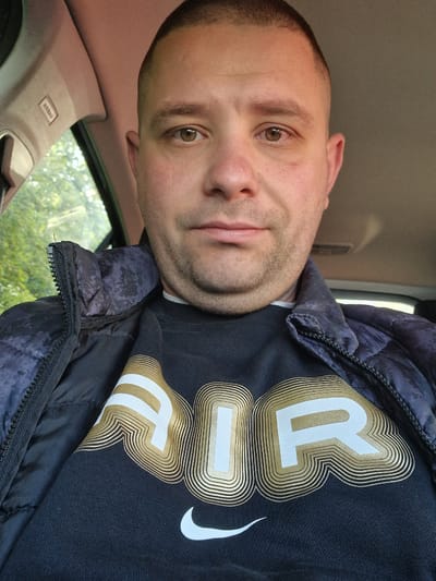 A medium shot captures a man inside a vehicle. He is wearing a black t-shirt with "AIR" written in white with gold accents, and a dark jacket. The interior of the car is visible, with an "AIRBAG" label on a panel above him. Outside the car window, a blurry view of foliage suggests an outdoor setting. There's no explicit evidence to pinpoint the location to Bearpark, United Kingdom in this image. The shot focuses mainly on the man and the interior of the vehicle.