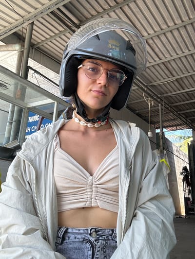 A young woman, wearing a gray helmet with a clear visor and a tan, ruched crop top, and a light-colored jacket, poses for a selfie under a corrugated metal roof structure. She has on glasses and a shell necklace. The background suggests an outdoor, urban setting with some construction or industrial elements, possibly in Hong Son, Vietnam, indicated by Vietnamese text on a sign. Sunlight filters through the structure.
