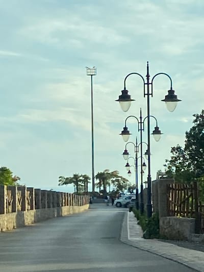 An asphalt road curves to the right, bordered on the left by a stone wall topped with decorative wooden posts. On the right, a sidewalk is separated from the road by a stone wall and a rustic wooden fence. Several ornate street lamps, each with multiple lights, line the right side of the road, casting long shadows. In the distance, palm trees and other foliage are visible. Two white vehicles are parked on the right side of the road, and a few people are walking near them. A tall, industrial-looking light pole stands in the background on the left. The sky is overcast with scattered clouds.