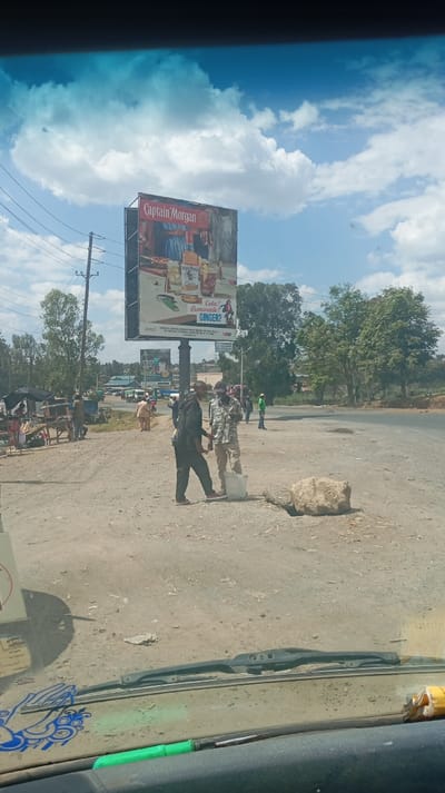 A roadside scene with two men standing near a large rock. In the background, there's a billboard advertising Captain Morgan rum. The area appears to be a small town or rural setting with trees and buildings visible. The photo is taken from inside a vehicle, with the dashboard and windshield visible in the foreground. The weather is sunny with some clouds in the sky.