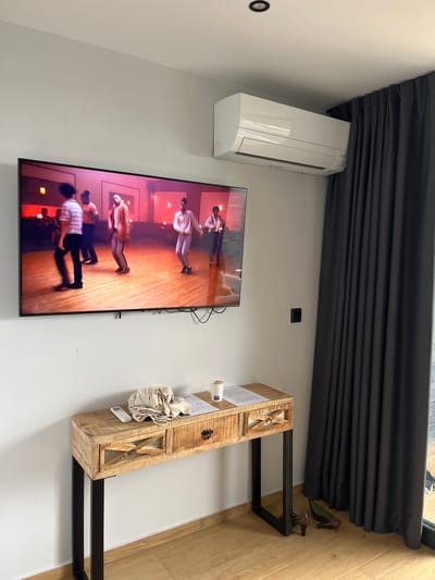 The image depicts a room with a television mounted on the wall, an air conditioning unit above it, and a console table below. The television displays a scene of people dancing in a room with a red glow. On the console table, there is a remote control, a folded cloth, a cup of coffee, and some papers. To the right of the console table, dark grey curtains are drawn, and a pair of high-heeled shoes are on the wooden floor. The room has white walls and a light wooden floor. The overall impression is a modern and somewhat minimalist interior. This scene is situated within the city of Can Güell, Spain.