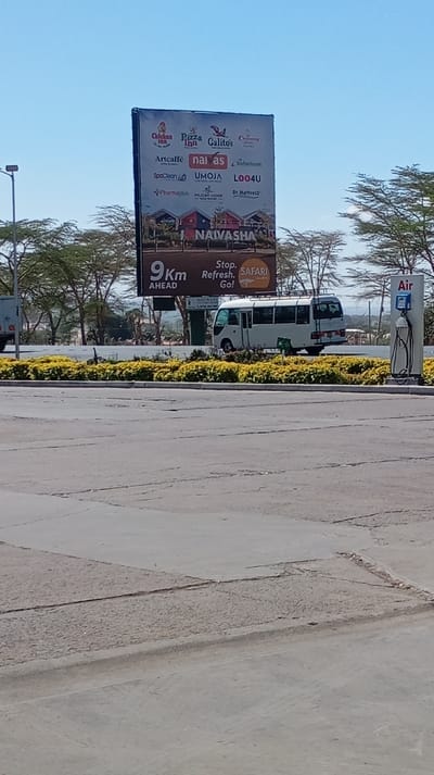 The image captures a sunny, daytime scene featuring a large roadside billboard, a paved area, and some vegetation, suggesting a location along a travel route.
In the center of the frame, a prominent, tall rectangular billboard dominates the midground. It features advertisements for various businesses and services, laid out in a grid pattern. Readable names include "Chicken Inn," "Pizza Inn," "Galito's," "Artcaffe," "naivas," "Safaricom," "SpaClean," "UMOJA," "L004U," "Pharmaplus," "PELICAN LODGE," and "Dr. Mattress." Below these logos, the word "NAIVASHA" is displayed in large letters, accompanied by small illustrations resembling houses or lodges. At the bottom left of the billboard, the text "9Km AHEAD" is visible, while on the bottom right, it reads "Stop. Refresh. Go!" next to a circular graphic with the word "SAFARI" and an animal illustration (possibly an elephant or rhino).
The foreground consists of a wide, grey paved surface, likely part of a road, parking lot, or service station, showing some cracks and wear. A low, well-maintained line of green bushes with yellow flowers separates the paved foreground from the area behind it.
Behind these bushes, a white minibus, typical of a public transport vehicle (possibly a matatu), is parked or driving, facing slightly away from the camera. It has a roof rack. To the far left, a portion of a truck is visible. On the far right, a vertical white structure with a hose, labeled "Air," indicates an air pump station, commonly found at fuel stations or service areas.
In the background, beyond the immediate roadside elements, there are numerous trees with sparse foliage, typical of a savanna or dry landscape, under a clear, bright blue sky. The overall impression is that of a commercial stop along a highway in a warm, possibly African, region, given the explicit mention of "Naivasha" and "Safari" which are associated with Kenya.