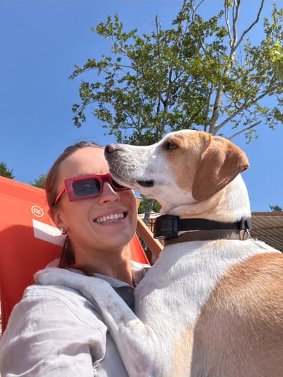 A young woman with brown hair pulled back, wearing red-framed sunglasses and a light-colored shirt, is seated in an orange chair outdoors. She is smiling, and a tan and white dog is leaning in, its nose touching her face. The dog wears a dark collar with a black device attached. Behind them, green trees and a clear blue sky are visible. The background suggests an outdoor recreational area, possibly near a restaurant or cafe with thatched roofing structures visible. The overall scene is one of relaxed interaction between a person and her pet on a sunny day. The location is identified as Kuk, Montenegro.