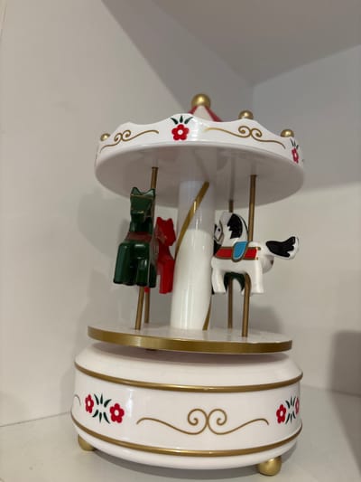 A close-up, eye-level shot shows a small, white carousel music box decorated with gold trim and red flowers. The music box has three visible carousel animals: a dark green horse, a red horse, and a white horse with black markings. The music box is situated on a white shelf, likely indoors. There are no people or any other objects visible in the frame.