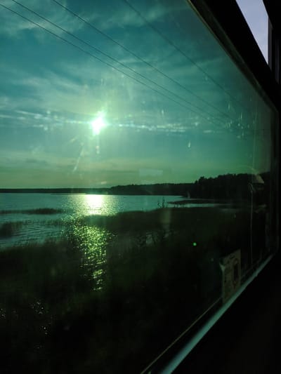 This image captures a tranquil view of a body of water and its surroundings, likely taken from a moving vehicle, possibly a train, due to the perspective and the reflection of power lines on the window. The sun is setting or rising, casting a bright glare on the water and creating a shimmering effect. The sky is a mix of blue and wispy clouds, tinged with a greenish hue, possibly due to the window's tint or atmospheric conditions.
In the foreground, dark vegetation lines the edge of the water. The water itself appears calm, with ripples and reflections of the sky and sunlight. In the distance, a dense line of trees forms the horizon. The overall impression is one of natural beauty and serenity.
The location of Osel'ki, Russia, is mentioned. Osel'ki is a settlement located in the Vyborgsky District of Leningrad Oblast, situated on the northern shore of the Gulf of Finland. The imagery of water, trees, and a potentially vast sky is consistent with a location in this region of Russia. Without further information or context, it is not possible to identify specific landmarks or features within Osel'ki from this image alone.