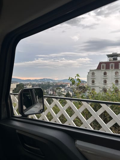 The image captures a view from inside a vehicle looking out the window towards Lam Vien - Da Lat, Vietnam. The foreground features the interior of a car, with a black car door and a side mirror visible. The side mirror reflects the interior of the car and a person wearing a striped shirt. Beyond the car's window, a white lattice fence runs horizontally across the frame. In the mid-ground, a large white building with a red roof and arched windows stands prominently. This building appears to be a hotel or a similar establishment. The background reveals a city skyline with numerous buildings, nestled amongst rolling hills. The sky is overcast with a mix of gray and white clouds, with hints of orange and pink suggesting it might be dawn or dusk. The overall scene depicts an urban landscape viewed from a vehicle, with a particular focus on the architecture and the natural surroundings of Da Lat.