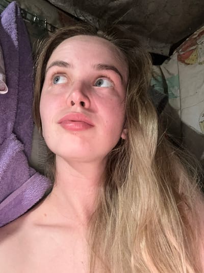 A close-up shot features a young woman with blonde hair, looking upwards and to her left. She is lying down, and a portion of a purple towel or robe is visible on the left side of the frame. The background is dark and appears to be an indoor setting with some indistinct patterns on what might be bedding or upholstery on the right. The lighting is dim, casting shadows. The location is identified as Азинский, Russia.