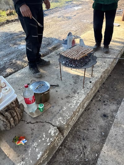 The scene depicts an outdoor cooking event in Palamartsa, Bulgaria, under natural daylight. Two individuals are partially visible, standing on a cracked concrete surface. The person on the left wears black pants with reflective strips and dark work boots, holding a silver fork in their right hand. The person on the right wears dark pants and shoes, with a green garment partially visible.
A portable charcoal grill occupies the center of the frame, laden with approximately 17-18 raw or lightly cooked sausages (likely kebapcheta or similar) arranged on its metal grate over glowing charcoal. Behind the grill, a red and white leaf blower is positioned, presumably for fanning the coals.
To the left of the grill, on the concrete, a large plastic bottle of Coca-Cola with a red cap rests on a piece of cardboard, next to a small green container and a silver-colored metal pot with a lid. Further left, a white plastic container holding several white mushrooms is placed on a section of a tree trunk. A crumpled, multicolored piece of trash lies on the concrete in the foreground.
The immediate ground consists of worn, cracked concrete. Beyond the concrete, a dirt path or road with visible tire tracks extends into the background, where sparse green vegetation is present. Light smoke emanates from the grill.