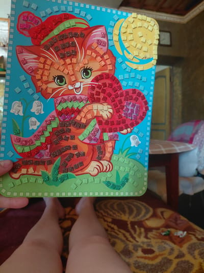 An image captures a woman's legs and a mosaic craft depicting a cat holding a heart, positioned indoors. The craft is likely a children's activity kit, characterized by its colorful glittery mosaic tiles forming the image of a ginger cat with a red hat, a green and red striped scarf, and a red heart. The background reveals an indoor setting with a patterned rug and a glimpse of furniture. The specific location of Marynivka, Ukraine, is not discernible from the visual information presented in the image.