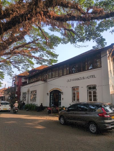 The image depicts a street scene in Fort Kochi, Kochi, Kerala, India. The prominent structure is a white-walled building identified by text as the "Old Harbour Hotel." The hotel features a large arched entrance in the center, flanked by rectangular windows, and a continuous band of windows on the upper floor, all beneath a tiled roof.
Overhead, large tree branches covered in epiphytes and moss extend across the frame against a clear blue sky.
On the paved road in front of the hotel, a dark grey multi-purpose vehicle (likely a Maruti Suzuki Ertiga based on visible features) is parked on the right side, facing away from the viewer. Its license plate begins with "KL," indicating Kerala registration. Further down the street, a white hatchback (Hyundai i10 model or similar) is visible. A person on a light-colored scooter or motorcycle, wearing a light-colored shirt and helmet, is traveling away from the camera in the left lane.
Several individuals are present near the hotel entrance and on the adjacent sidewalk. Some are seated on steps or benches outside the hotel, while others appear to be standing. Two scooters or motorcycles are parked near the hotel entrance steps. The general environment suggests an established urban area with trees providing natural canopy.