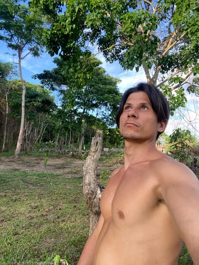A shirtless man with dark hair and brown eyes is standing outdoors in what appears to be a wooded area. He is looking upwards and to the right, with his head tilted slightly back. The man's torso is visible, showing his chest and shoulder. Behind him, there are several trees with green leaves, and a blue sky with some white clouds is visible through the branches. The ground is covered in grass and dirt, with some fallen leaves. In the background, there is more dense vegetation and trees, suggesting a natural or rural setting. The lighting indicates it is daytime, likely with sunlight filtering through the trees. No specific landmarks or structures that would definitively place this in Los Velásquez, Venezuela, are visible.