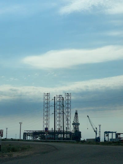 The image depicts an industrial landscape under a cloudy blue sky. Dominating the center are two tall, lattice-like structures, likely part of an oil rig or industrial facility. To the right of these, a more prominent drilling rig structure is visible, alongside a large crane. In the foreground, a paved road curves towards the right, with dry, sparse vegetation bordering it. Several utility poles and power lines crisscross the scene, attesting to the industrial nature of the location. A circular sign with the number "35" is affixed to a pole near the road. The overall impression is one of a functional, working industrial zone, likely situated in or near the city of Aktau, Kazakhstan, a region known for its oil and gas industry. There are no people visible in the frame.