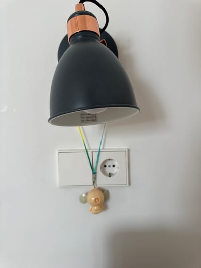 A black wall-mounted lamp with a copper accent is pictured hanging above a white electrical outlet on a white wall. A lanyard with a small, light-colored cartoon charm hangs from the lamp and dangles near the outlet. The outlet appears to be a standard European socket. The image does not provide any information or context that would place it in Murcia, Spain.