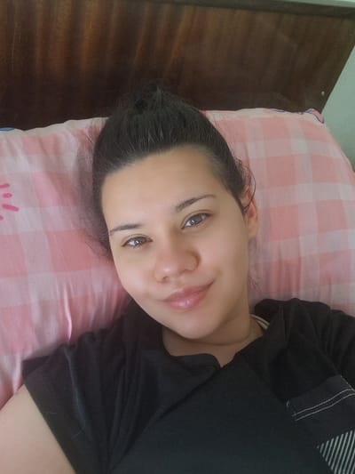 A young woman with dark hair pulled up in a bun is lying on her side on a pink and white checkered pillow. She is wearing a black t-shirt. Her head is slightly tilted, and she is looking at the camera with a gentle smile. The background appears to be a wooden headboard. There is no discernible city or geographical context that would indicate Raduvene, Bulgaria.