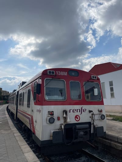 A stationary Renfe commuter train, identified by the marking "138M" on its upper red facade and the "renfe" logo in purple on its white lower front, is positioned on railway tracks at what is identified as the Buñol train station in Buñol, Spain. The train's front features two rectangular windshields, a central coupling apparatus, horns, and various light fixtures; the white section of its exterior displays visible dirt and wear. The train rests alongside a light-colored, tiled platform. To the left, the tracks curve into the distance, paralleled by an elevated embankment where obscured structures are faintly visible. To the right, a white building with a distinct red-pitched roof and several rectangular windows stands. The scene is outdoors, illuminated by daylight under a sky with significant coverage by large, scattered grey clouds, interspersed with areas of clear blue sky. No individuals are present in the frame, and no actions or interactions are depicted.
