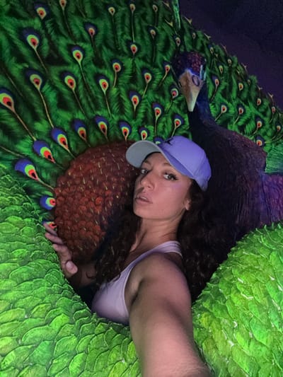 A person wearing a light purple baseball cap and a light purple tank top is lying on what appears to be a large, artificial peacock. The person has brown curly hair and is looking at the camera. The peacock has green and brown feathers with detailed eye-like patterns. The scene appears to be a staged photo opportunity, potentially in a tourist attraction. The image's location is Binh Lu, Vietnam.