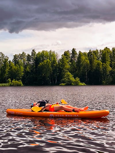 In Vecumnieki, Latvia, a person is lying supine on an orange inflatable paddleboard on a body of water. The person is wearing orange shorts, a black vest, and blue goggles. A yellow paddle is resting on the board next to the person's torso, with another paddle visible at the head of the board. The paddleboard has "FUSION 10'10"x32"" and "AQUA MARINA" printed on it. The water is dark and rippling. In the background, a line of lush green trees borders the water, with a cloudy sky overhead.