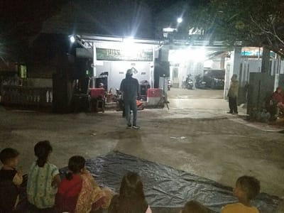 An outdoor night gathering is captured in Baleendah, Indonesia, with a group of people facing a brightly lit stage-like area in front of buildings. In the foreground, several children are seated on a dark tarp spread on the concrete ground, their backs to the viewer, appearing to watch an activity.

In the mid-ground, a man in a dark jacket and jeans stands facing the lit area, with sound equipment, including large speakers and a red cooler, positioned around him. Another man in a light-colored shirt stands further right, near the buildings. The background features several brightly illuminated buildings, possibly residential or community structures, with multiple artificial lights creating significant glare. Parked motorcycles are visible in front of these buildings, and some foliage frames the scene from the right.

The visible text on a banner or sign on the main building reads "PUSKESM", likely indicating a "Puskesmas" or community health center, suggesting the event might be related to public health or community welfare. The scene depicts an informal community event or gathering, with participants focused on the central illuminated area, possibly for a performance, presentation, or screening. The overall atmosphere suggests a communal night activity.