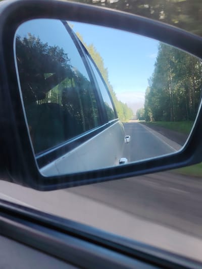 The image depicts a view from a car's side mirror while driving on a road. The mirror reflects a straight asphalt road stretching into the distance, flanked by green trees on both sides. The sky visible above the road is light blue. A portion of the car the mirror is attached to is also visible, showing its side and part of the window frame. The picture seems to be taken while the car is in motion. The location might be in the vicinity of Гремяча, Russia.