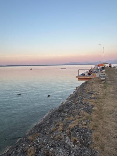 The image depicts a tranquil evening scene at a lake, likely Chiemsee, given the location context of Gstadt am Chiemsee, Germany. A stone embankment runs along the right side of the frame, transitioning into a grassy area. A wooden pier juts out into the calm water, occupied by several people lounging and relaxing. Two umbrellas, one orange and one yellow, provide shade. A sailboat mast is visible in the background. Two ducks are swimming in the foreground of the water. The sky displays a gradient of soft blues and pinks, indicative of sunset or twilight, with distant mountains silhouetted against the horizon. The water is clear near the shore and deepens in color further out. The overall atmosphere is serene and relaxed, suggesting a popular spot for leisure.