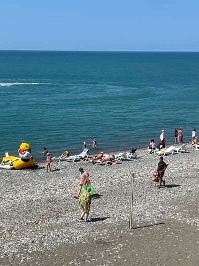 The image shows a crowded beach in Giachrypsh, Abkhazia. The sea is a bright turquoise color and appears calm. The beach is covered in grey pebbles and there are several people enjoying the sun and sea. In the foreground, a person in a yellow and green patterned outfit walks with a green inflatable ring. Behind them, a person in a pink t-shirt and red shorts walks with a bag.
Further back, there are people sunbathing on lounge chairs and wading in the water. To the left, a large inflatable yellow duck-like toy is partially submerged in the water. There are also several beach umbrellas and chairs scattered across the pebbles. In the background, the sky is a clear blue. The scene depicts a typical summer day at the beach with various recreational activities taking place.