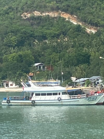 A landscape-oriented, daytime photograph captures a scene in Baan Chaloklum, Thailand. A multi-story boat is situated in the water along the shoreline. The boat's name "Sail Rock Divers" is visible. The boat is docked next to buildings with slanted roofing. Lush, green, dense forest covers the left side of the photograph. The water appears murky.