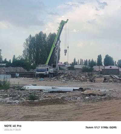 The image depicts a construction site in Sergeli, Uzbekistan, under a cloudy sky. A large green crane with the brand "ZOOMILION" is prominently featured, with its boom extended. The crane is positioned over a construction area with piles of debris, wood, and building materials, including some long metal sheets in the foreground. A white truck, likely a cement mixer or similar construction vehicle, is parked near the crane. In the background, residential buildings with pitched roofs are visible behind a fence, surrounded by tall trees. The ground is uneven, with sand, rocks, and dirt. The overall scene suggests ongoing construction or demolition work. The timestamp "2026.06.17 06:33" and camera details "NOTE 40 Pro 74mm f/1.7 1/198s ISO95" are overlaid on the image.