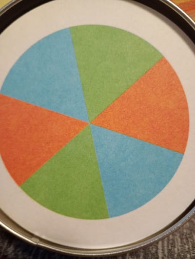 The image shows a close-up, overhead view of a round object with a pie chart design. The pie chart is divided into six equal segments of alternating blue, green, and orange colors. The object appears to be a lid or a spinning wheel, made of white paper or cardstock, set within a metallic rim. The background is dark and out of focus, suggesting a casual indoor setting. There are no people, animals, or specific landmarks visible in the image. The location in Rosenheim, Germany, is not identifiable from the visual information provided.