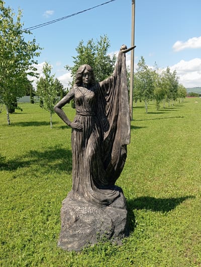 A full shot captures a bronze statue of a woman standing on a rock base in a grassy, sunlit field. The statue depicts a female figure with flowing drapery, her left hand on her hip and her right arm extended upwards, holding a small object. The base of the statue is inscribed with Cyrillic lettering, appearing to read "ДЕВА".
The surrounding environment consists of a well-maintained lawn, with scattered trees and a line of birch trees receding into the background. A utility pole stands to the right of the statue, and power lines are visible across the bright blue sky, which is dotted with fluffy white clouds. The overall scene suggests an outdoor public space in Otradnaya, Russia, possibly a park or a cultural landmark.