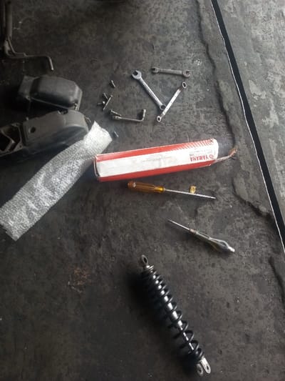 An assortment of tools and motorcycle components rests on a soiled, dark gray surface. A black shock absorber lies diagonally at the bottom. A yellow-handled screwdriver and another with a transparent handle are centrally located near a red and white Yamaha parts box. Wrenches of varying sizes are scattered near a disassembled engine cover and bubble wrap. Various nuts, bolts, and a bent metal tool are also present.