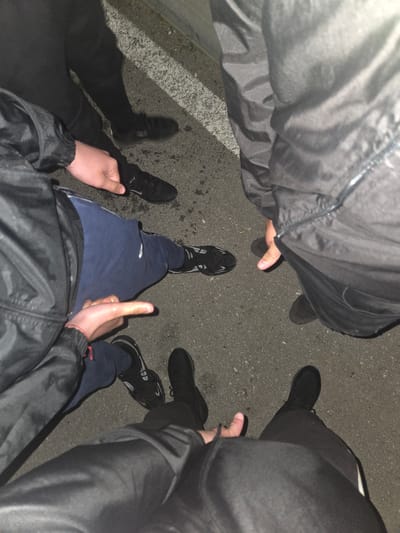 Four individuals stand in a circle on asphalt, their feet and lower bodies visible. The surface shows signs of wear and possible staining. All four are wearing dark-colored shoes or sneakers. One person is wearing blue sweatpants. The individuals are also wearing dark jackets. The scene suggests an outdoor setting, but the presence of a white line on the lower left suggests an urbanized area. The image contains insufficient data to link to the location specified.
