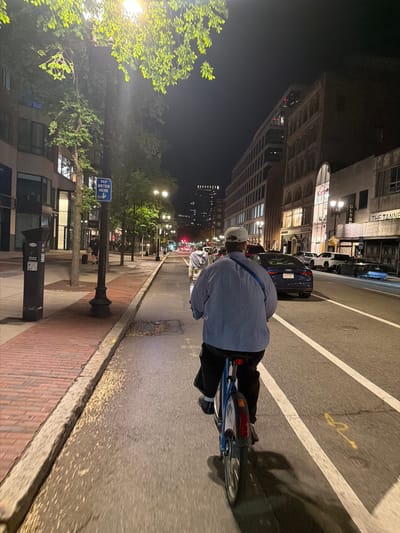 At night, the image depicts a man riding a blue bicycle on a city street in Boston, Massachusetts. He wears a light-colored baseball cap and a blue striped shirt. To his left is a brick sidewalk, a streetlamp, and a building bearing the sign "PAY METER HERE." Across the street are taller buildings and trees, all illuminated by streetlights. Several cars are visible in the adjacent lane. The street is divided by painted white lines.