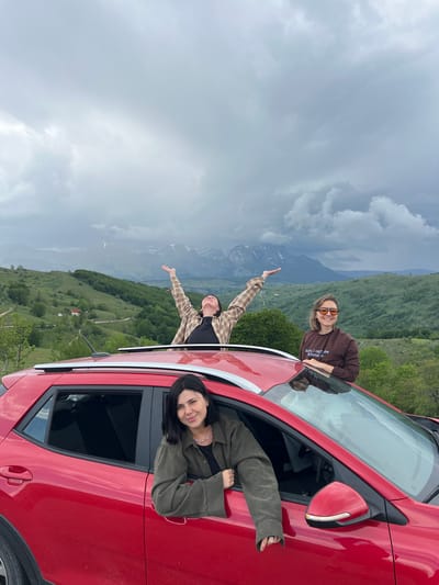 Three women are posing by a red car on a hillside in Pišče, Montenegro. Two are sticking their heads and arms out of the vehicle, while another woman leans on top of the car. The surrounding environment includes mountainous terrain and a cloudy sky.