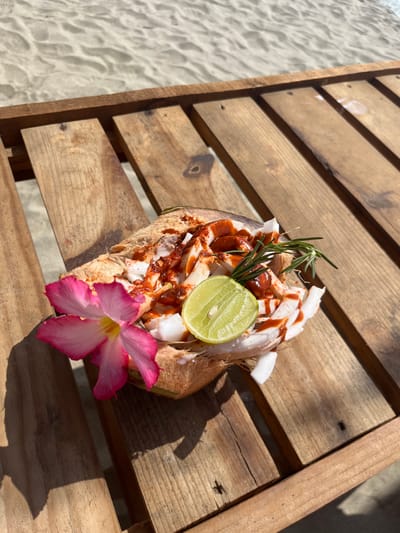 A close-up shot shows a halved coconut filled with coconut meat, topped with a red sauce, a lime wedge, and a sprig of rosemary. The coconut is adorned with a pink and white desert rose flower at its base. It rests on a weathered wooden deck, with sand visible in the background, suggesting a beach setting. The presence of sand, the style of the deck, and the type of flower suggest this may be in Brisas de Zicatela, Mexico.