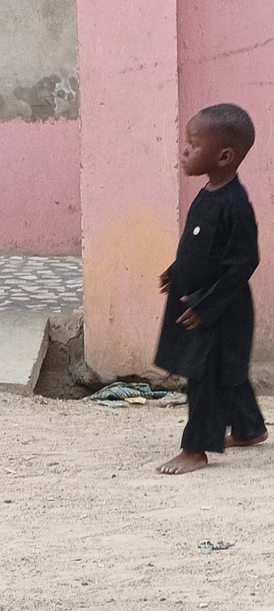 An outdoor scene in Rangaza, Nigeria, features a young, dark-skinned child captured in a profile view, facing towards the left side of the frame. The child is dressed in a traditional-style black long-sleeved tunic and matching trousers, with a small, circular white badge pinned to the left side of their chest. They are barefoot, walking or standing on a sandy, dusty ground speckled with small pebbles and debris.
The background consists of a textured wall, primarily painted pink, with patches where the paint appears faded or chipped, revealing lighter, possibly gray, sections underneath. To the child's left, a rough, gray concrete structure, possibly a step or foundation, is visible at the base of the wall. Near this structure and on the ground lies a crumpled piece of blue and white striped fabric.
The lighting indicates it is daytime. The child's expression is calm and focused, gazing ahead. No legible text is visible on the badge or elsewhere in the image.