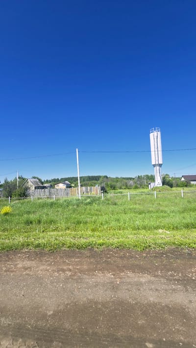 The image captures a rural scene under a clear blue sky. In the foreground, a dirt road runs horizontally across the bottom of the frame. Beyond the road, a strip of lush green grass extends towards a weathered wooden fence. Behind the fence, several houses with wooden fences and roofs are visible amidst trees and vegetation. To the far right of the scene, a tall, cylindrical white water tower stands prominently against the sky. Power lines are strung across the middle ground. The overall impression is that of a quiet, rural settlement. The location is identified as Актюбинский, Russia.