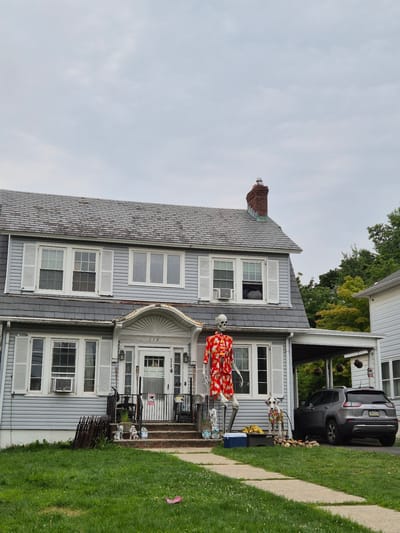 This media file captures the exterior of a two-story house in South River, United States of America. The house is light blue with white shutters on its windows. A dark grey SUV is parked in the driveway. The house is decorated with several skeletal figures, one of which is dressed in a bright red and yellow floral outfit. There are also smaller figures, possibly gnomes or other decorative items, near the entrance. A black metal fence surrounds the porch area. The scene appears to be set up for a holiday, likely Halloween, given the presence of the skeletons. The weather is overcast, suggesting a cloudy day. The image is oriented vertically, showing the house from a slightly elevated perspective, with a lawn and a concrete path in the foreground. The house number "114" is visible above the front door.