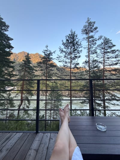 The image depicts a person's bare legs crossed as they recline on a dark wooden deck. The deck overlooks a wide, fast-flowing river and a densely forested mountainside, bathed in the warm light of either sunrise or sunset. Pine trees are prominent in the foreground and middle ground. A black metal railing separates the deck from the view of the river. On the deck, to the right of the person's legs, is a small, clear glass ashtray with an unlit cigarette. The scene appears to be in a natural, outdoor setting, likely in or near the city of Barangol, Russia, given the context. The sky is a clear, pale blue.
