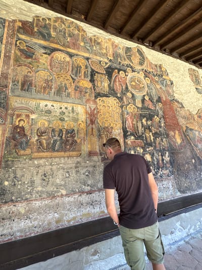 A male subject is depicted from behind, intently viewing a large, ancient outdoor fresco on a textured stone wall. The subject wears a dark brown short-sleeved t-shirt, olive green cargo shorts, and sunglasses are perched on his head. His hands are placed in his pockets as he leans forward towards the mural.
The fresco, stylistically characteristic of Byzantine or Orthodox iconography, covers a substantial portion of the wall, displaying numerous religious scenes and figures rendered in earthy tones of gold, dark blue, red, brown, and green. Notable compositions include a lower-left panel depicting a seated female figure and three seated male figures, all with halos, set against a verdant background with trees. Other sections feature celestial beings, architectural motifs, groups of saints enclosed within medallions, and narrative scenes.
The artwork exhibits considerable degradation consistent with age and environmental exposure, manifesting as significant paint loss, extensive cracking, and fading, particularly on its lower sections. The lower register also bears faint graffiti-like marks and a horizontal delineation of faded white and red paint residue. A dark-stained wooden beamed ceiling structure or overhang is visible directly above the fresco, suggesting a covered area. The ground surface beneath the subject is composed of irregular stone paving. This scene is situated at a religious edifice within the vicinity of Sandanski, Bulgaria.