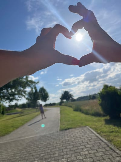 In Sachsenheim, Germany, the sun shines brightly through a heart shape formed by two hands, held up in the foreground. The sun's rays create a lens flare across the image. In the background, a paved path winds through a grassy area with trees and fields. A person, out of focus, walks away from the camera down the path. The sky is blue with scattered white clouds.