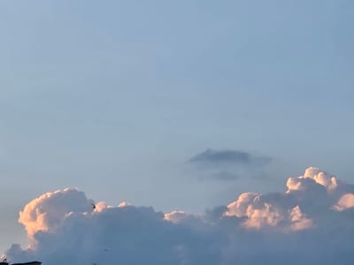 The image displays a serene sky over Širvintos, Lithuania, at either dawn or dusk. The sky is a soft, pale blue, transitioning to lighter hues towards the top. Dominating the lower half of the frame are large, fluffy cumulus clouds, tinged with warm, rosy light from the setting or rising sun. Some clouds are illuminated from within, creating a soft glow, while others are in shadow, appearing in shades of gray and white. A few smaller, wispy clouds are scattered higher in the sky. Two small, dark silhouettes of birds are captured in mid-flight, adding a dynamic element to the tranquil scene. In the bottom left corner, a sliver of a building's roofline is visible, suggesting an urban or suburban setting within Širvintos. The overall atmosphere is peaceful and picturesque.