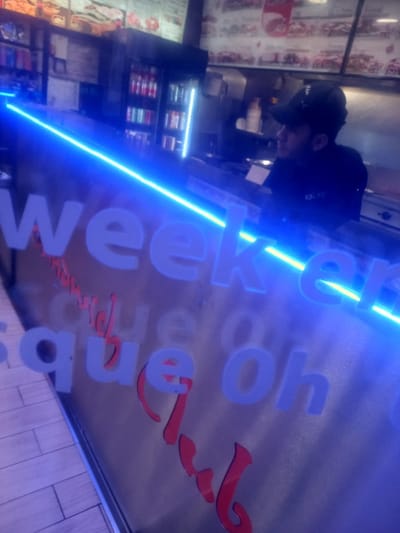 A person wearing a hat is behind a counter. Behind them, a menu board displays various food items. The counter has a sign with illuminated blue letters and a red embellishment, partially reading "Weeker." The lighting is dim and creates a blurry, dynamic effect, and the image is rotated.
