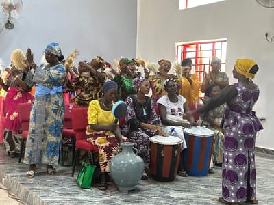 The image depicts an indoor gathering of approximately 18-20 adult women in Goni Gara, Nigeria, engaged in a communal activity, likely a performance or worship service. The setting is a brightly lit room with plain light-colored walls and a patterned gray tiled floor, possibly a community hall or place of worship, featuring red-framed windows with security bars on the right and a white wall-mounted oscillating fan on the left.
The women are dressed in vibrant, traditional African patterned attire, primarily long skirts or dresses and matching head wraps in various colors including blue, green, yellow, purple, and brown. Several women on the left and in the middle are standing, clapping their hands, and holding whisk-like objects, indicating active participation in a dance or ritual. Their expressions appear engaged.
On the right side of the image, four women are seated on red upholstered chairs, forming a musical ensemble. One woman in a yellow top plays a blue and black maraca-like instrument. Two other women, dressed in dark patterned and white embroidered tops respectively, are playing two large, cylindrical drums (one red and black striped, the other red and blue striped). A fourth seated woman is partially visible. In the foreground, on the floor, is a large, gray, unglazed pottery vase with two handles, and a green tote bag.
Standing on the far right, facing the seated musicians and other participants, is a woman in a purple and white patterned dress and a yellow head wrap, with her right arm extended, appearing to lead or direct the group. The overall scene suggests a vibrant cultural or religious event involving music, dance, and collective participation.