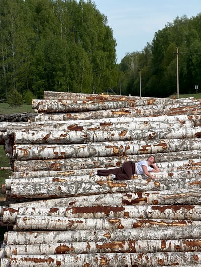 The image depicts a young woman lying on a stack of birch logs in a rural setting. The woman is wearing a white t-shirt, brown sweatpants, and tennis shoes. She is positioned lengthwise along the logs, with her head resting on her arm.
The logs are stacked high and appear to be recently cut, based on their clean, white bark. The background shows a forest landscape with green trees and a clear sky.
Based on the location provided, the scene is likely located in or near Ural'skoe, Russia.