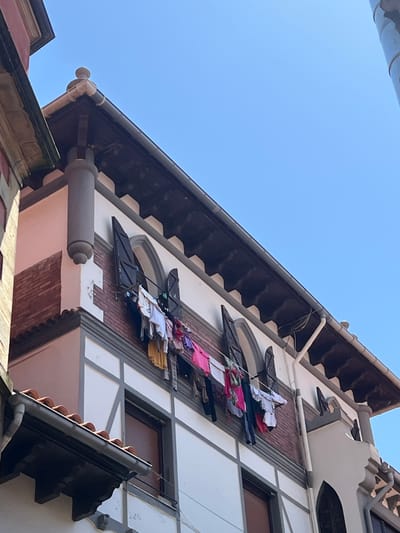A low-angle, medium shot captures a portion of a building in San Sebastián, Spain, with laundry hanging out to dry. The building features white stucco walls with dark gray decorative trim and wooden beams. Brickwork is visible behind two arched windows on the second story, each fitted with dark shutters that are partially open. Laundry, including t-shirts, pants, and towels in various colors, hangs on a clothesline strung across the windows. The roofline is dark brown with decorative eaves. Above, a clear blue sky dominates the upper portion of the frame. The architecture suggests a traditional European style.