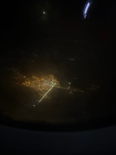 A night-time view from an airplane window reveals a city's lights below. Blurred by the glass and altitude, the cityscape appears as a constellation of golden dots, concentrated around a central cluster. A road or bridge stretches out from the main area, marked by a line of lights. The dark expanse of the plane interior and the window frame occupy the edges of the image. A rectangular light source is visible above.