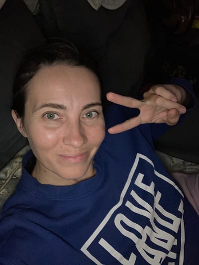 A woman lies indoors, looking directly at the camera with a serene expression, her head tilted slightly. She has fair skin, dark hair, and striking green eyes, which are wide open and engaged. A faint smile plays on her lips, suggesting a calm or content mood. Her right arm is raised, forming a peace sign (V-sign) with her index and middle fingers.
She is wearing a dark blue, long-sleeved sweatshirt featuring a prominent, stylized white text on the chest that clearly spells out "LOVE," enclosed within a bold white rectangular outline. The background is softly lit and consists of dark, textured fabrics, likely a couch or bedding, with a lighter, patterned fabric visible behind her head. The dim lighting suggests the photograph was taken during the evening or night. The overall scene conveys a relaxed, comfortable atmosphere within a private indoor setting, situated in Florianópolis, Brazil.