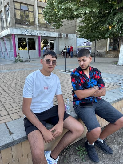 Two young men are sitting on the edge of a brick sidewalk. The man in the foreground wears a white t-shirt and sunglasses. The other man sits behind him and wears a floral shirt. They are in front of a building with a sign in Cyrillic script that says "General." There is a tree to the left of the building. A poster is visible on a pole. People are seated at tables in the background. The location is identified as Hayredin, Bulgaria.