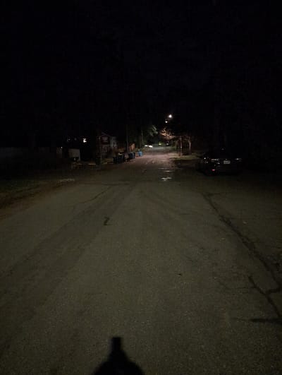 A dimly lit street stretches into the distance on the right side of the image. Buildings and sparse streetlights are visible on the left, receding into darkness. A parked car is visible in the center of the street. The asphalt road is cracked and appears wet in spots. A dark, blurry shadow is visible on the bottom right corner. The image is rotated and has poor lighting.